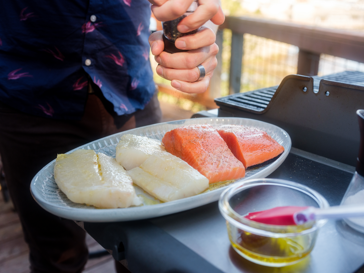 Outdoor grilling preparation with wild Alaskan salmon and halibut fillets being seasoned with pepper, fresh sustainable seafood from Bristol Bay fishermen