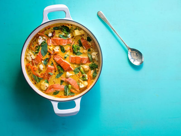 Wild-caught Alaskan salmon coconut curry with vegetables in a white pot on a turquoise background. A vibrant, healthy seafood dish made with sustainable Alaskan salmon.
