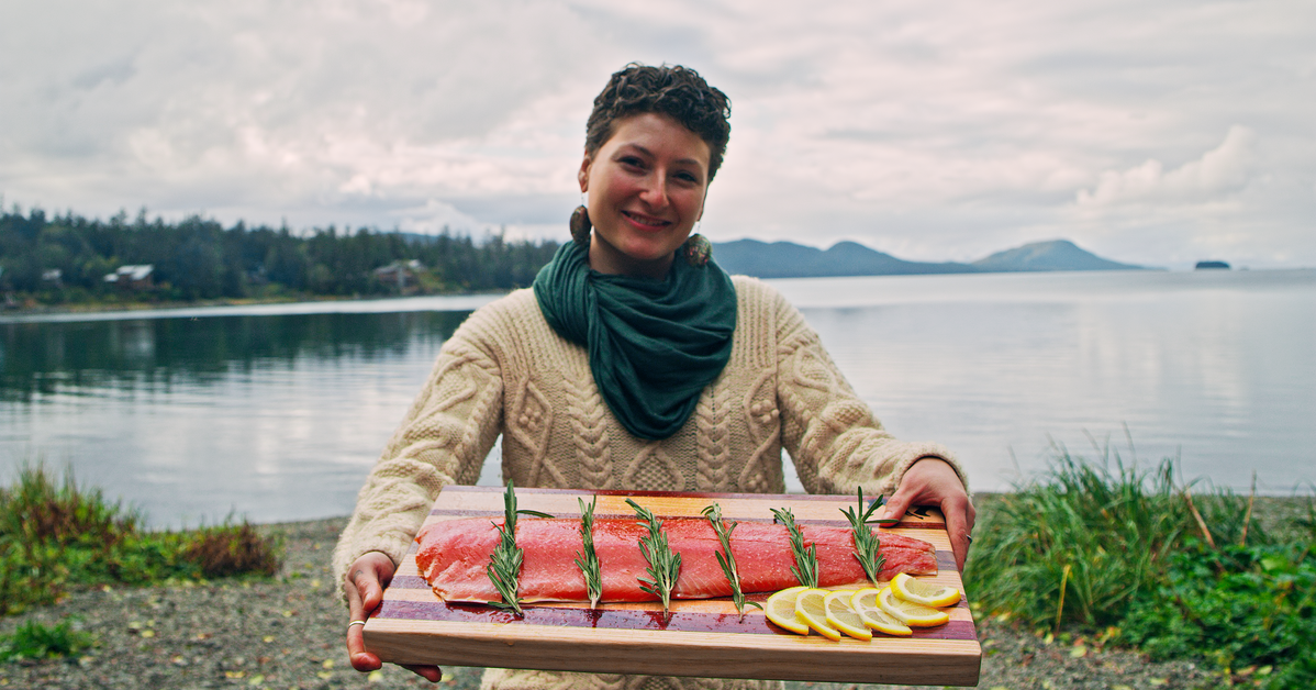 Person holding a fresh wild-caught Alaskan sockeye salmon fillet garnished with rosemary and lemon by the water in coastal Alaska. Sustainably harvested seafood from Net to Table Seafoods.
