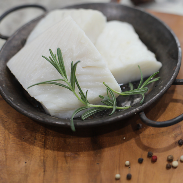Raw fillets of wild-caught Alaskan Halibut in a rustic metal dish garnished with fresh rosemary, showcasing the fish’s delicate white texture and premium quality.
