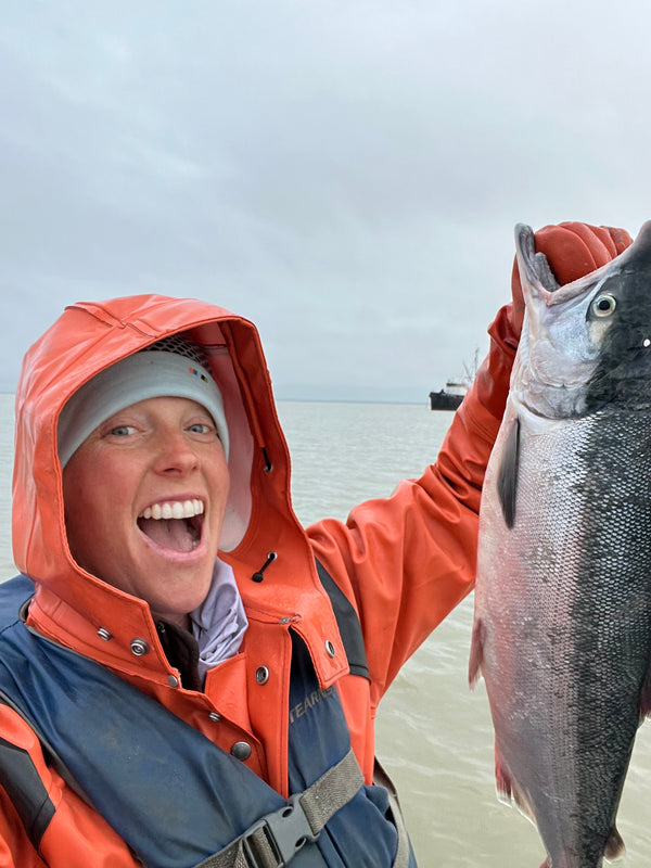 Smiling person in orange rain gear holding up a freshly caught Alaskan salmon near the water on an overcast day.
