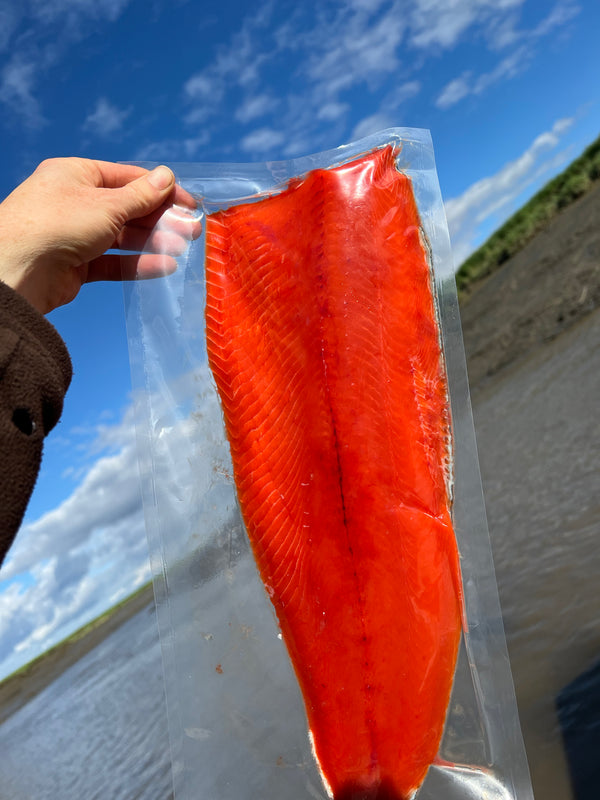 Hand holding a vacuum-sealed fillet of bright red wild-caught Alaskan sockeye salmon against a blue sky and river backdrop.
