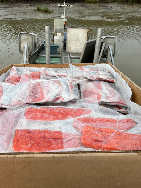 Box filled with vacuum-sealed wild-caught Alaskan salmon fillets on the deck of a small fishing boat docked in a muddy river channel.
