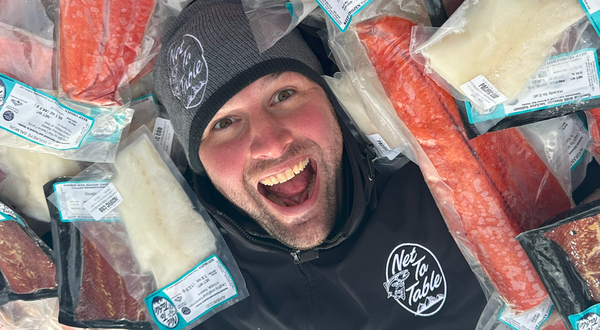 Captain Luke smiling man wearing a Net to Table Seafoods beanie and jacket surrounded by vacuum-sealed packages of wild-caught Alaskan fish, including salmon, cod, and halibut.
