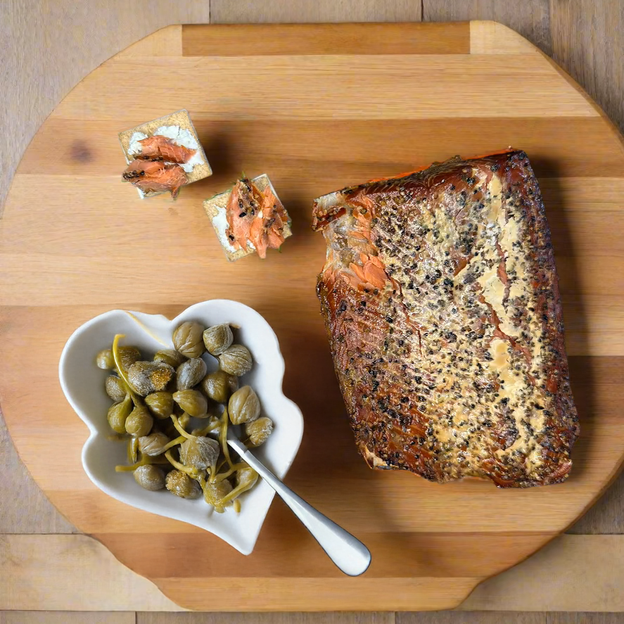 Herb-crusted smoked wild Alaskan salmon fillet on wooden cutting board with capers in white bowl