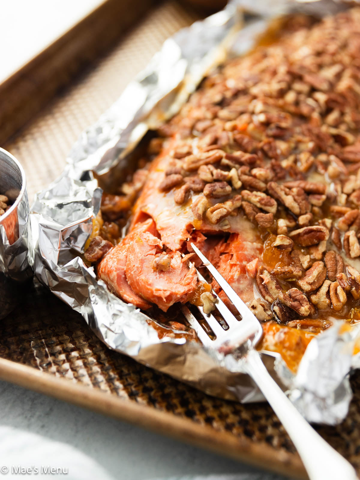 Baked wild Alaskan salmon fillet topped with chopped pecans and glaze, served on foil with a fork breaking into the tender fish.

