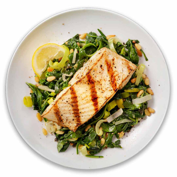 Grilled wild Alaskan halibut fillet served on a bed of sautéed spinach, leeks, and pine nuts with a lemon wedge garnish, showcasing a healthy and sustainable seafood meal.
