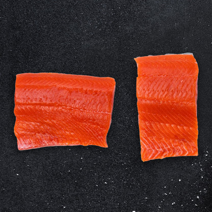 Two fillets of Wild Alaskan Sockeye Salmon from Net to Table Seafoods displayed on a dark stone background. Fresh, vibrant red wild-caught salmon sustainably harvested in Bristol Bay, Alaska.
